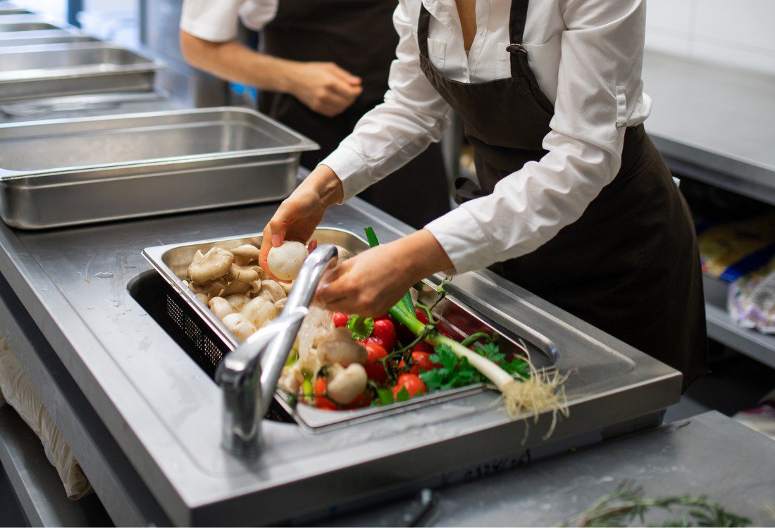A woman washing vegetables in one of the stainless steel sinks of the restaurant kitchen.