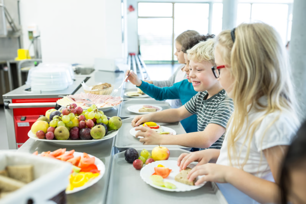 Children choosing fruit and breakfast items from a school kitchen serving counter with commercial catering equipment.