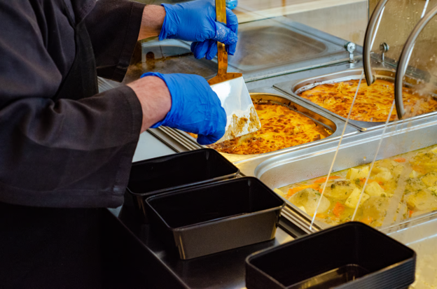 Commercial bain marie food counter with hot dishes being portioned into takeaway containers by a gloved catering worker.