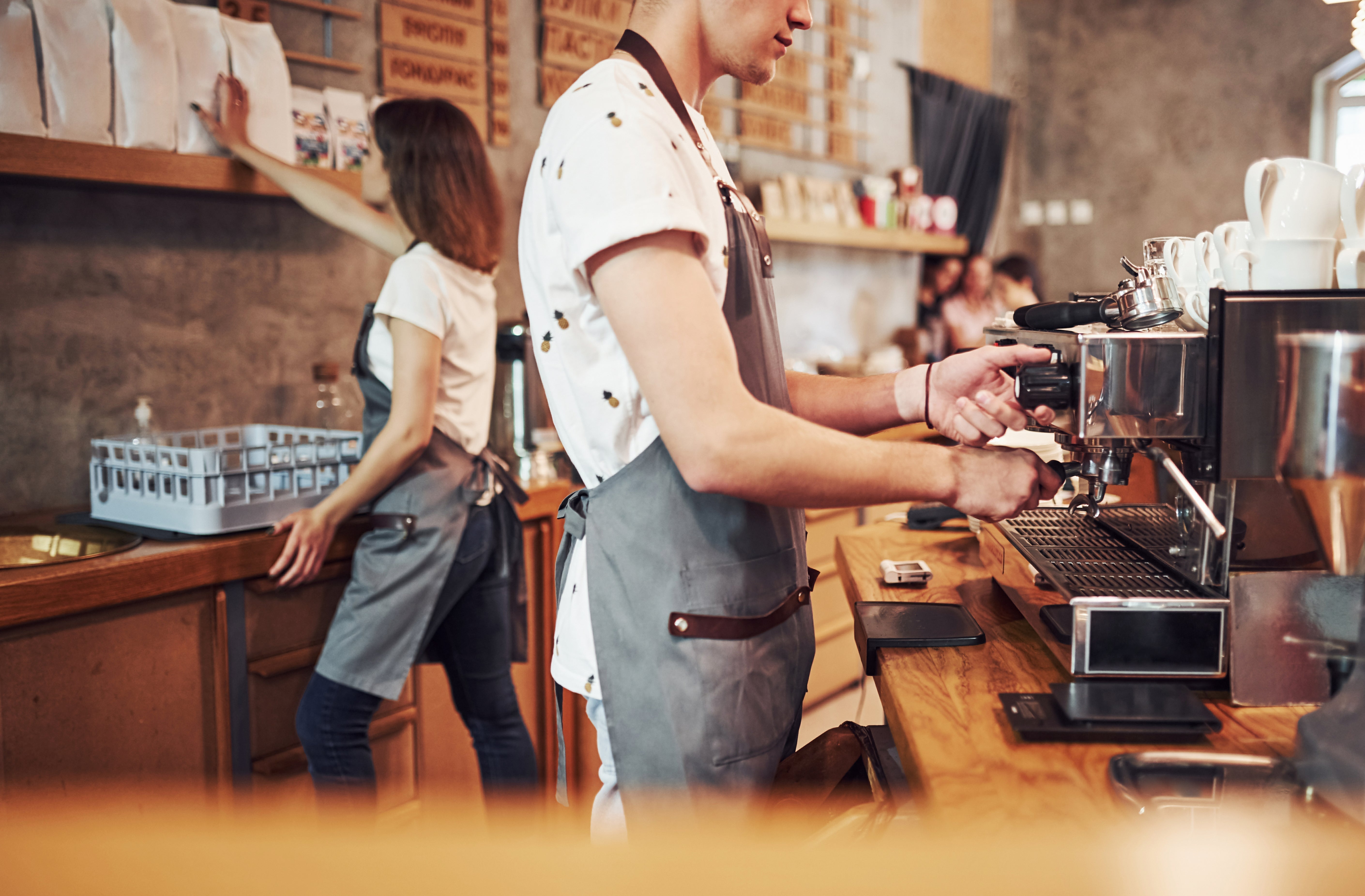 Barista preparing coffee with espresso machine in small coffee shop commercial kitchen setup.